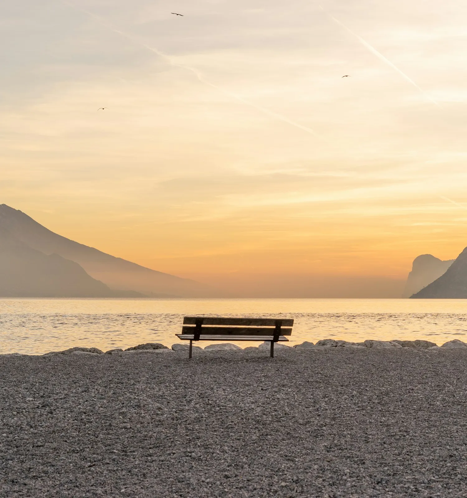 An empty bench on a pebble shore facing a lake, with mountains on both sides under a warm sunset sky