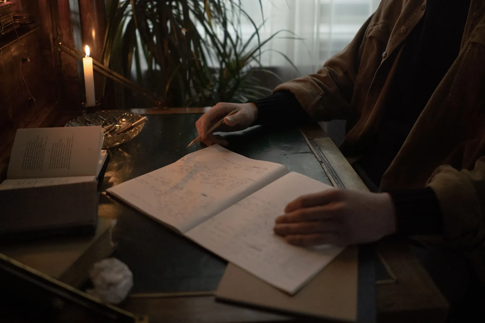 A person writing in an open notebook at a dark desk, with a lit candle, books and a plant behind