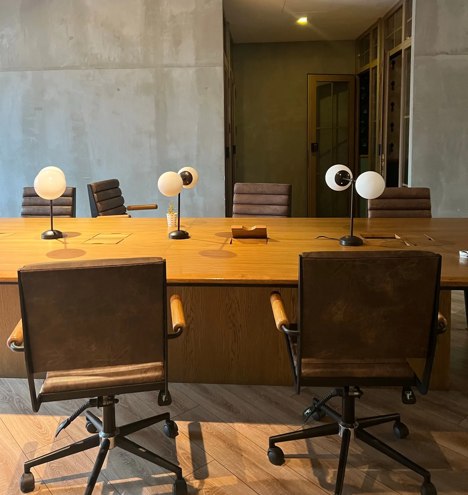 Long wooden meeting table with brown leather chairs and globe lamps in a room with concrete walls.