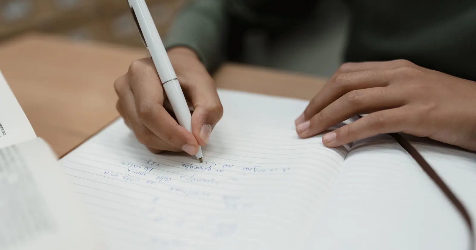 Close-up of hands writing with a white pen in a lined notebook on a light wooden table.
