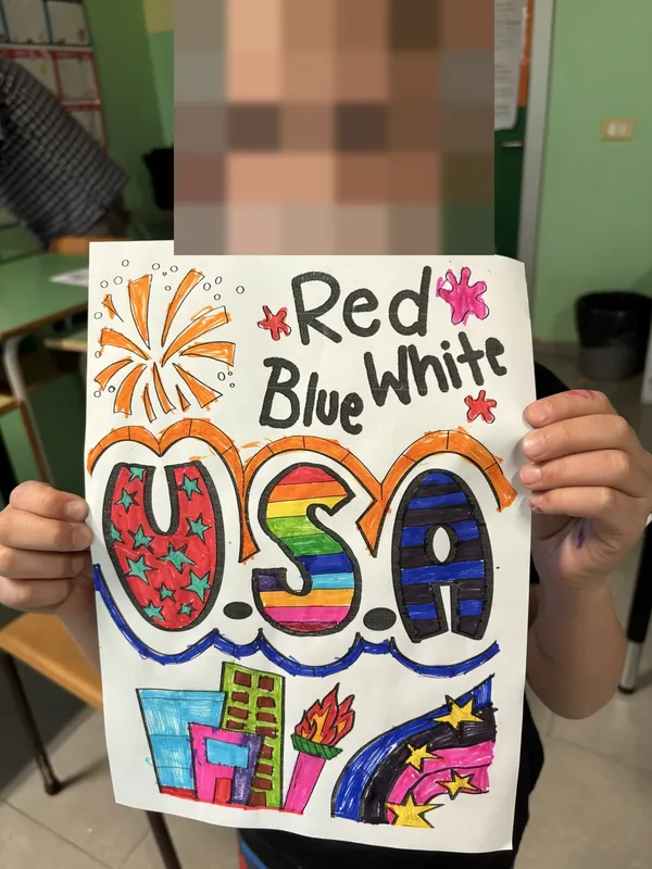 A boy holding up his colourful "Red Blue White USA" poster in a classroom.